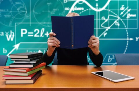 Ein Student neben einem Bücherstapel und einem Tablet vor einer Tafel mit mathematischen Formeln